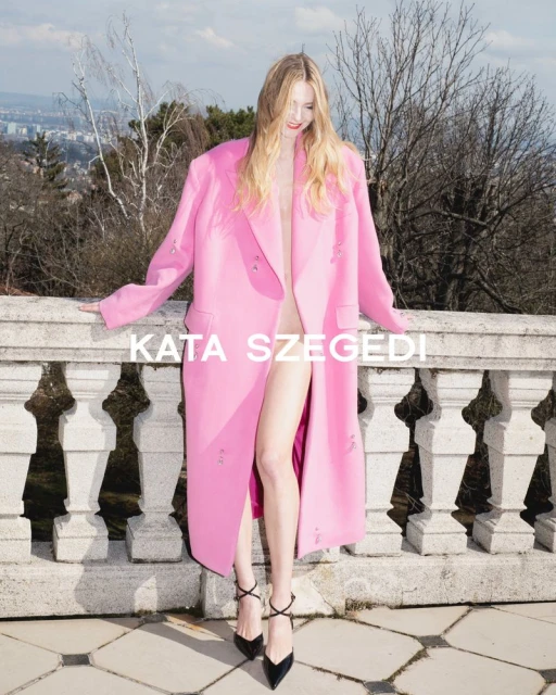 Fashion editorial shot of a fair-skinned adult woman posing in a pink coat and black heels on a stone balcony with a city view.