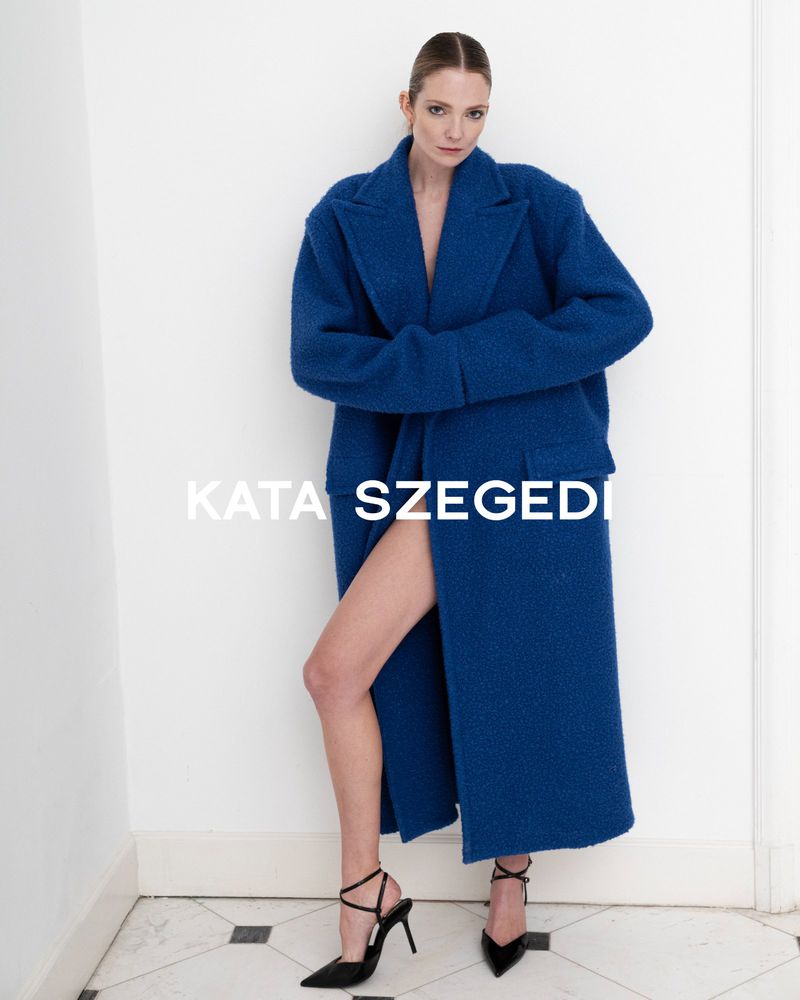 A fashion model poses indoors wearing a long, oversized royal blue coat and black high heels, standing against a white wall.