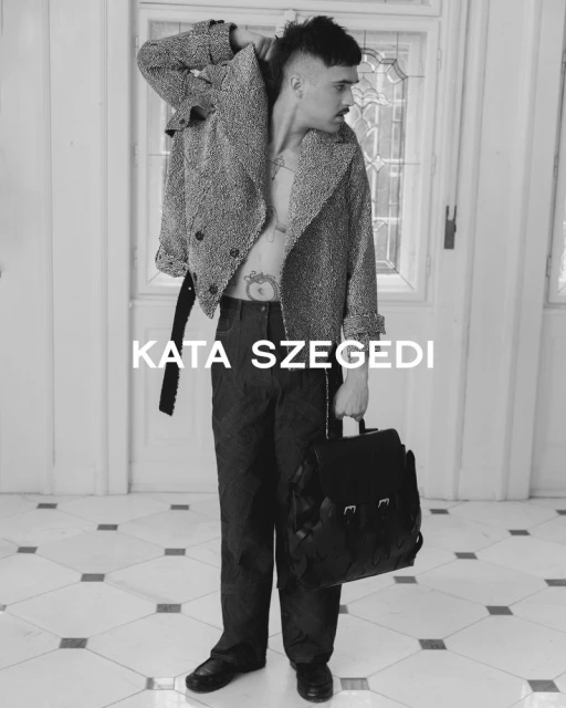 An adult male in a speckled jacket and dark trousers holds a bag in front of a white door and tiled floor, presented in black and white.