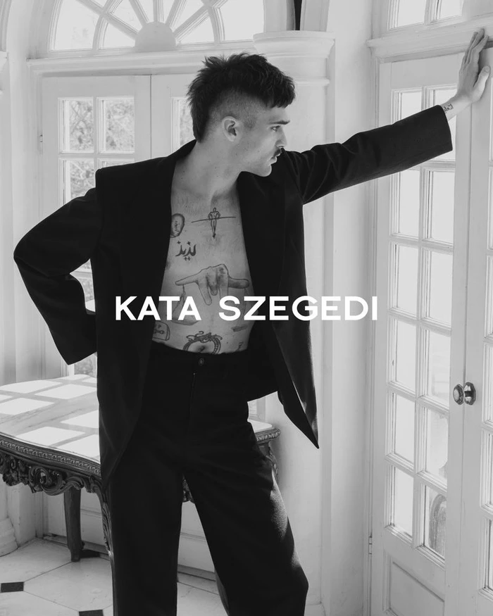 Monochrome fashion portrait of a tattooed man wearing a black suit jacket leaning against white panelled doors.