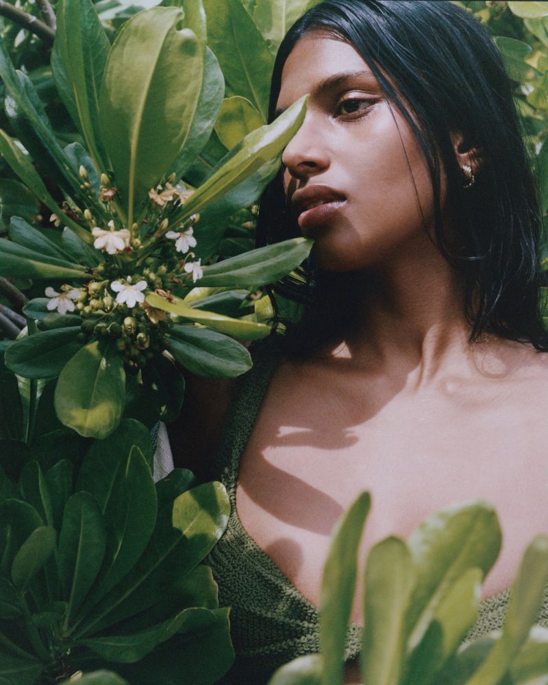 Close-up portrait of a woman with dark hair, light skin, and green top surrounded by tropical foliage and small white flowers.