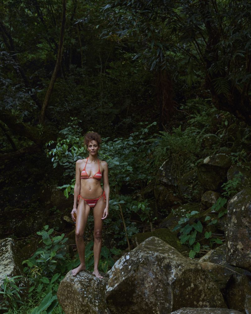 Woman in a striped bikini stands on a mossy rock in a lush, green forest setting. The natural light adds to the serene atmosphere.