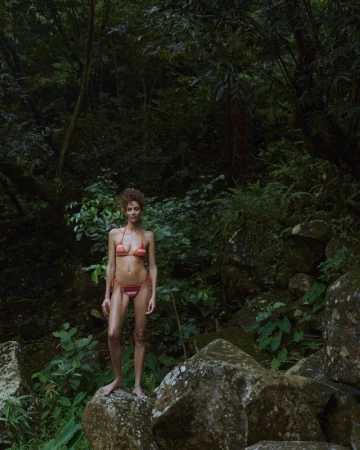 Woman in a striped bikini stands on a mossy rock in a lush, green forest setting. The natural light adds to the serene atmosphere.