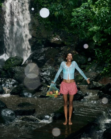 Woman stands on wooden bridge, holding fruit-filled bag, with waterfall and lush foliage. Wearing pink shorts and blue mesh top.