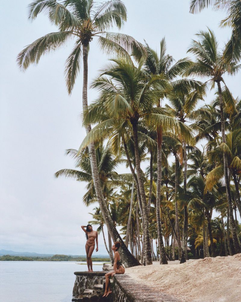 Two adult women pose on a stone pier near palm trees and calm ocean under an overcast sky, creating a tropical vacation scene.