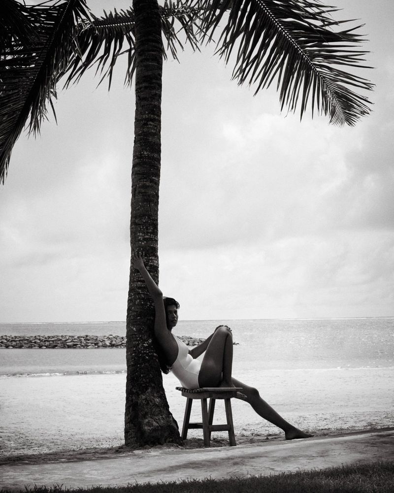 Model seated on a small wooden stool leaning against a palm tree trunk on a sandy beach with the ocean in the background