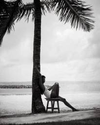 Model seated on a small wooden stool leaning against a palm tree trunk on a sandy beach with the ocean in the background