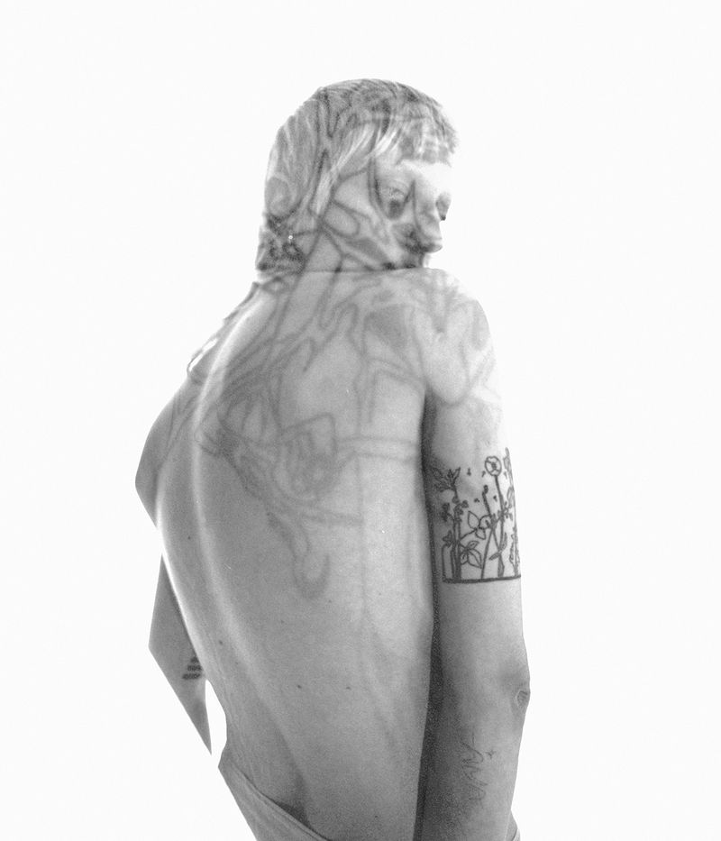 Monochrome portrait of a human with floral tattoo on the arm and abstract line art overlaid on the back and neck in a white space.