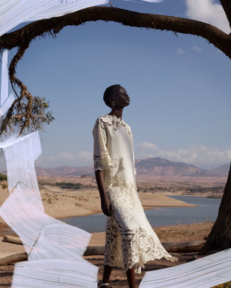 A dark-skinned woman in a white lace dress poses outdoors against a desert landscape under a large tree branch with white fabric.