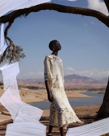 A dark-skinned woman in a white lace dress poses outdoors against a desert landscape under a large tree branch with white fabric.