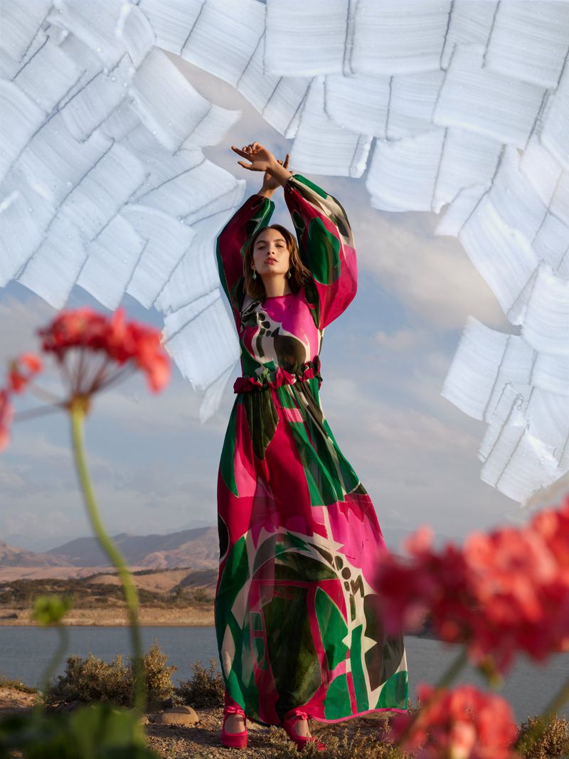 A woman in a long, flowing print dress stands outdoors with raised arms, framed by red flowers, against a water and mountain backdrop.
