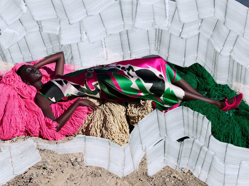 Woman in patterned dress lies on colorful yarn with a white geometric pattern, modeling fashion outdoors with bright, vivid tones.