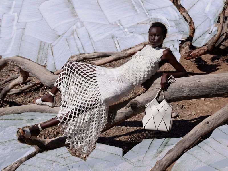 A dark-skinned woman in a white woven dress and sandals reclines on a fallen tree, carrying a white handbag, set against a landscape backdrop.