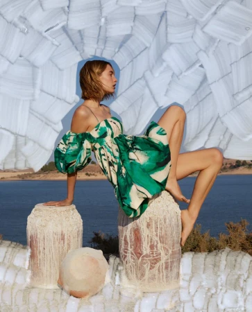 A fair-skinned woman in a green and white patterned dress poses on stone columns against a sea and sky backdrop under a modern art piece.