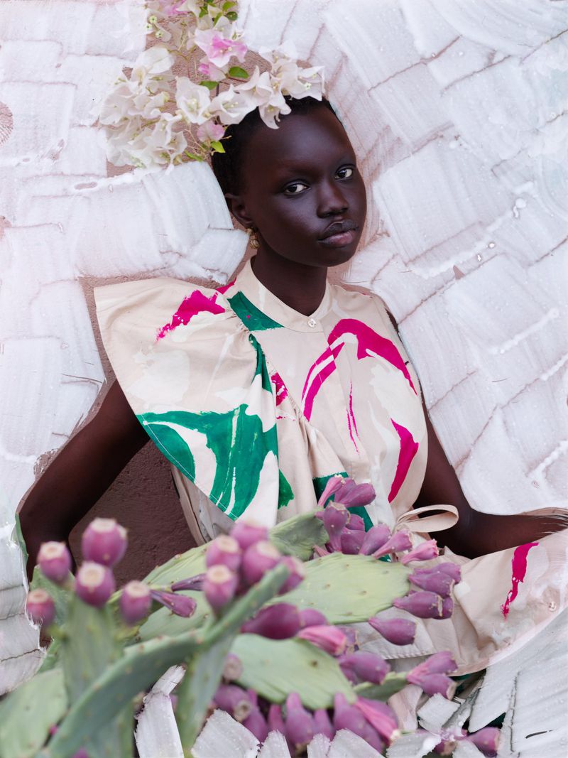 Portrait of a dark-skinned woman wearing a floral headpiece and a beige top with pink and green accents, posing in front of a textured wall.