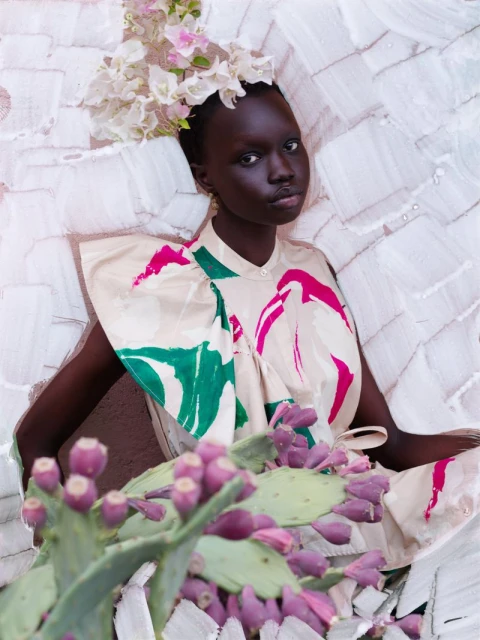 Portrait of a dark-skinned woman wearing a floral headpiece and a beige top with pink and green accents, posing in front of a textured wall.