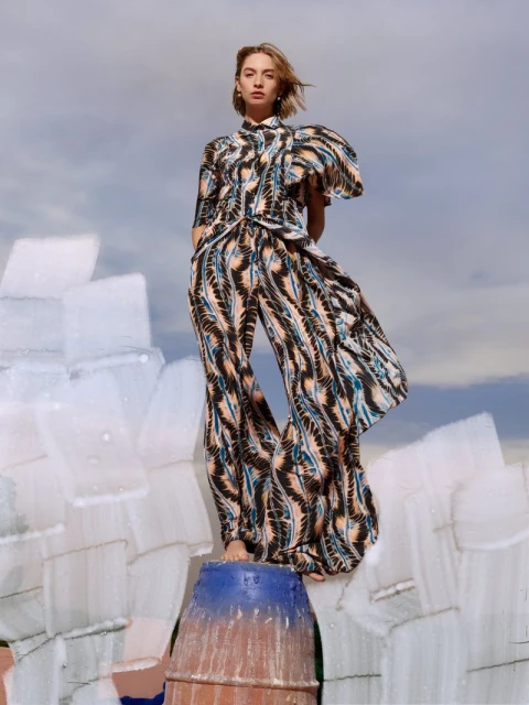 Woman in patterned co-ord set poses on textured structure against blue sky. Fashion editorial shot of model wearing a matching shirt and pants.