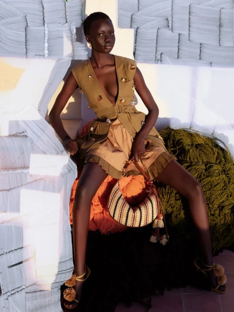 A dark-skinned woman models an olive green buttoned vest dress, accessorized with tasseled sandals and handbag, against a textured backdrop.