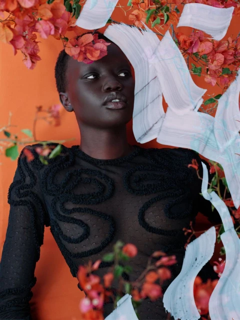 Portrait of a dark-skinned woman in a sheer black top, surrounded by orange flowers and a digitally painted background, looking off-camera.