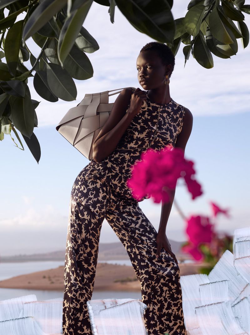 A dark-skinned woman in a patterned jumpsuit poses outdoors with a woven handbag, set against a backdrop of water and foliage.
