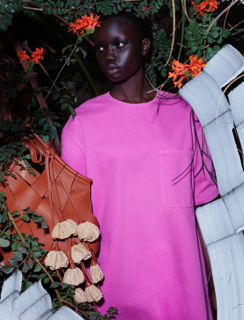 Portrait of a woman with dark skin wearing a pink dress, styled with a brown woven purse, amidst lush green foliage and orange flowers.