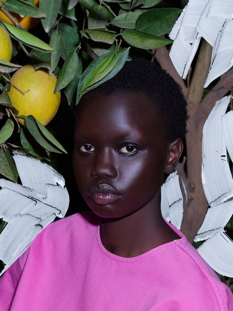 Headshot of a dark-skinned woman in a pink shirt, surrounded by greenery, branches, and yellow citrus fruits in a dark, intimate setting.
