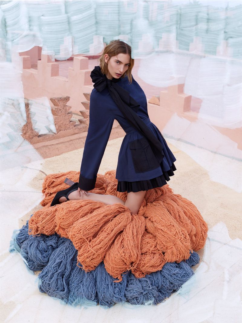 Adult female model kneeling on pile of orange and blue yarn, wearing blue jacket with a black ruffled collar, in a photo studio.