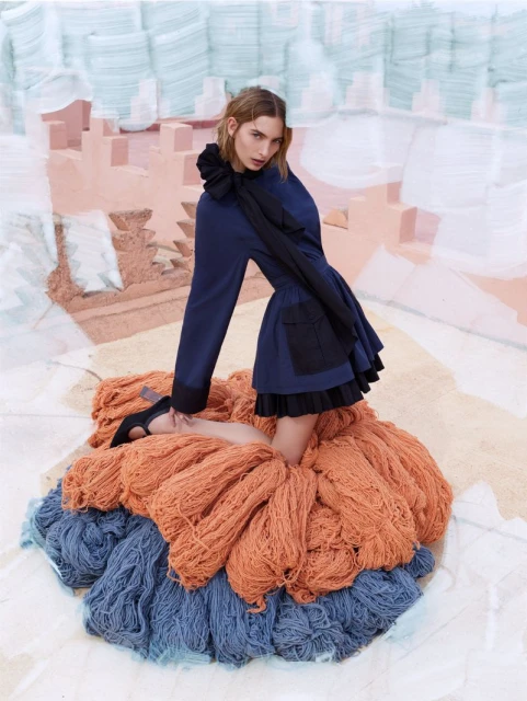 Adult female model kneeling on pile of orange and blue yarn, wearing blue jacket with a black ruffled collar, in a photo studio.