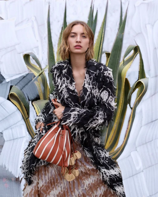 Fashion shot featuring a young adult woman in a textured coat and skirt, holding a striped bucket bag, against a background with agave plants.