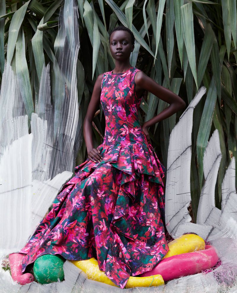 A South Sudanese woman poses outdoors, wearing a floral, sleeveless gown in front of lush green foliage and abstract white art.