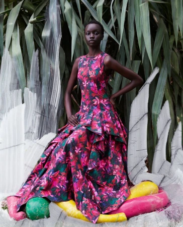 A South Sudanese woman poses outdoors, wearing a floral, sleeveless gown in front of lush green foliage and abstract white art.