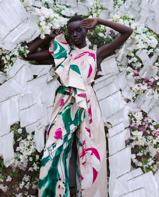 Elegant woman in a patterned jumpsuit poses with her hand near her face, laying among white flowers and textured white background, fashion editorial.