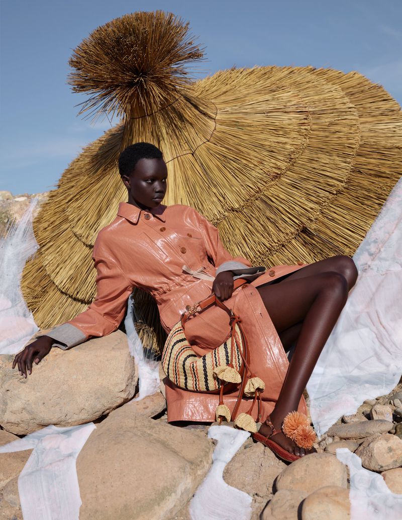 Fashionable woman poses with handbag and straw umbrella on rocks in a fashion editorial with clear sky background. Model wearing a peach coat.