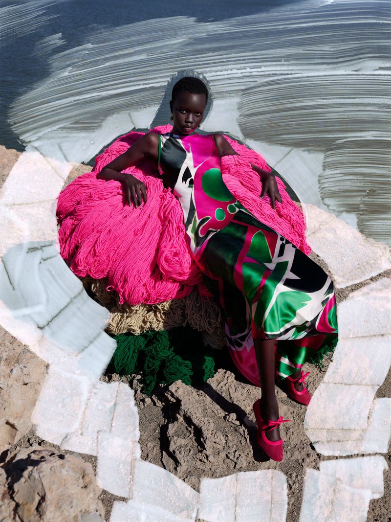A woman in a colorful patterned dress and pink shawl poses on rocks with a painted sky backdrop in a striking fashion photograph.