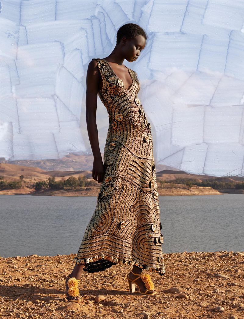 A dark-skinned woman models a crocheted gold gown with a V-neck and floral accents in a desert landscape by a lake under an overcast sky.