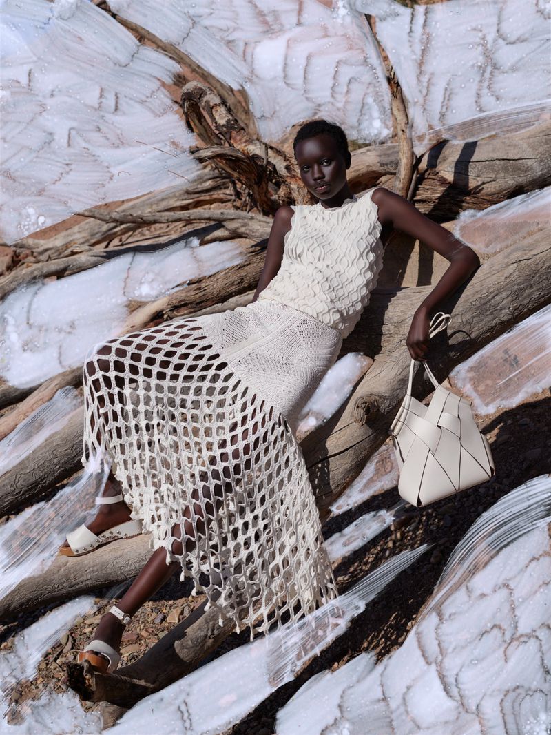 Fashion shot of a Black woman wearing a white knit dress sitting on driftwood with a tote bag in a bright, outdoor setting.