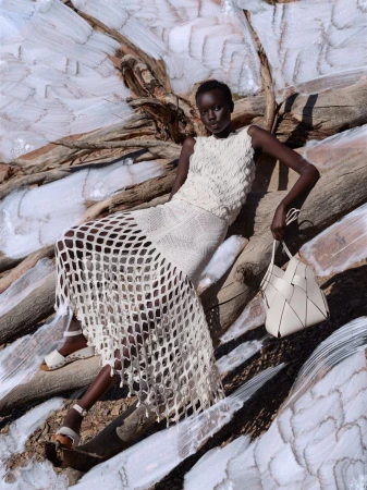 Fashion shot of a Black woman wearing a white knit dress sitting on driftwood with a tote bag in a bright, outdoor setting.