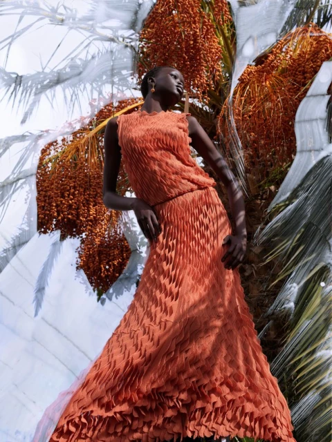 Woman in pleated orange gown poses with palm fronds and fruit. Fashion shot, full length, looking up and to the side.