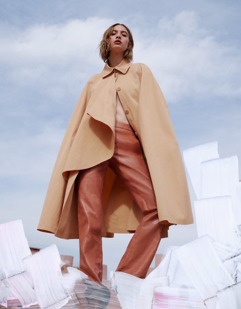 Fashion editorial shot of a blonde woman wearing a tan coat and rose colored pants posing on white geometric objects against a cloudy sky.