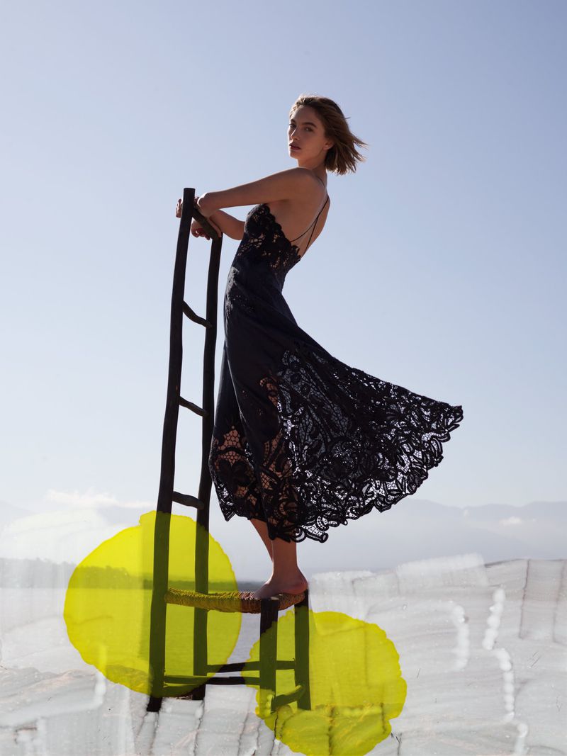 A woman in a black lace dress poses on a ladder in a light blue sky. An abstract yellow shape is in the foreground.