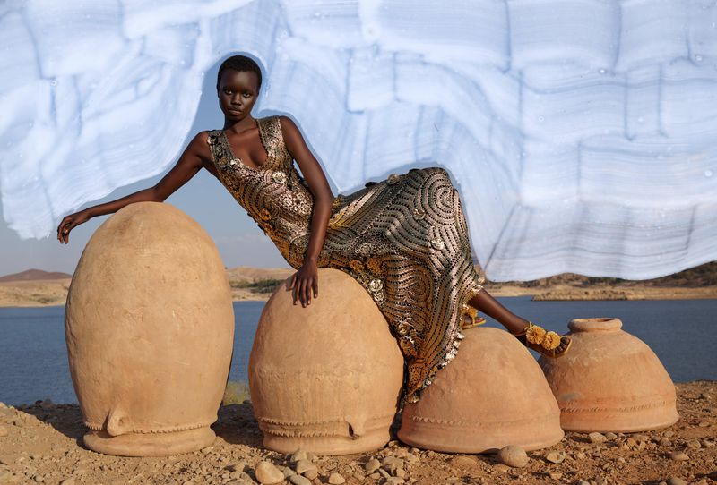 A regal African woman poses in an ornate dress while sitting atop a series of large, earthen pots in a serene landscape.