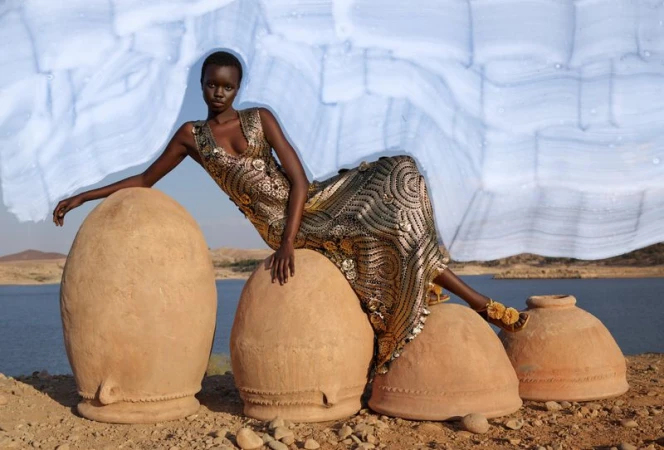 A regal African woman poses in an ornate dress while sitting atop a series of large, earthen pots in a serene landscape.