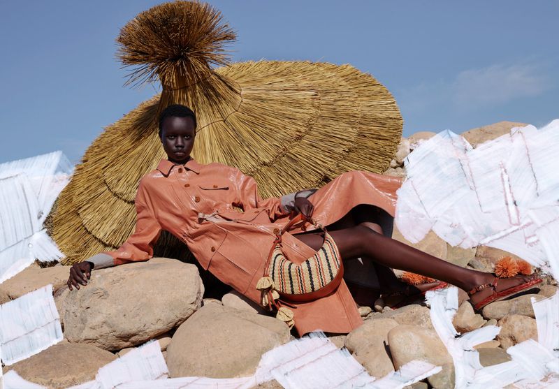 An African female model in a peach-colored dress reclines on rocks outdoors next to a straw umbrella and bag in a fashion shot.