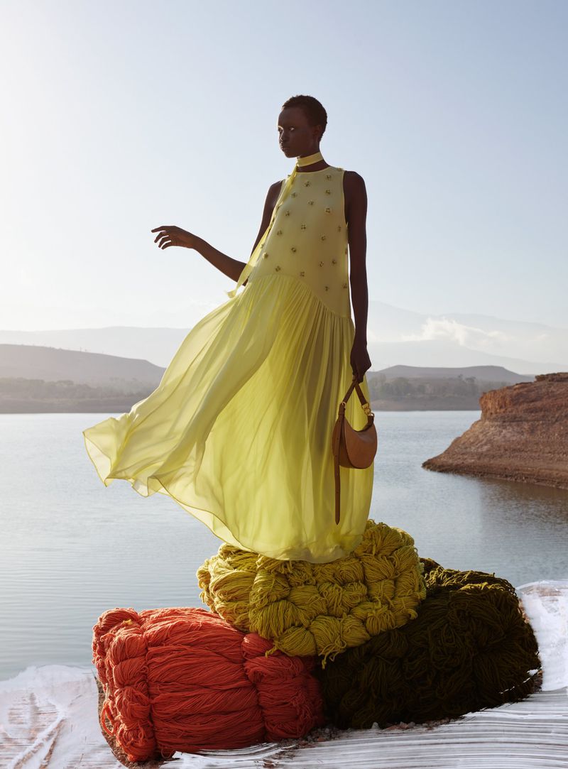An adult female fashion model poses in a flowing yellow gown on a stacked set of woven textile objects, against an outdoor lakeside backdrop.
