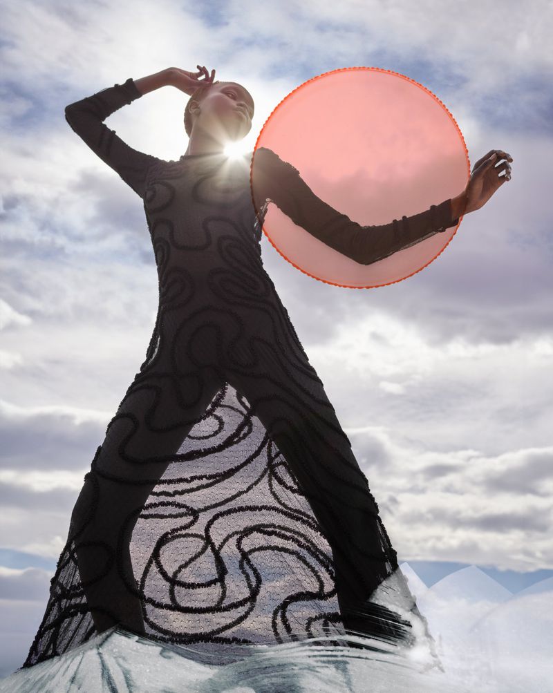 Fashion shot of a woman in a patterned black dress holding a circular pink object against a sky with clouds and bright sunlight.