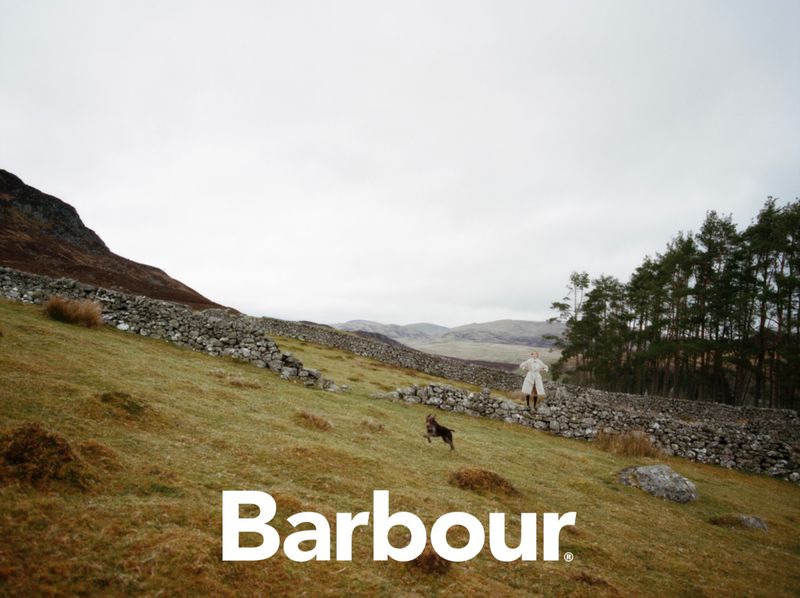 Outdoor shot of a woman in a coat and a dog running on a grassy hillside beneath a cloudy sky, featuring a stone wall and trees.