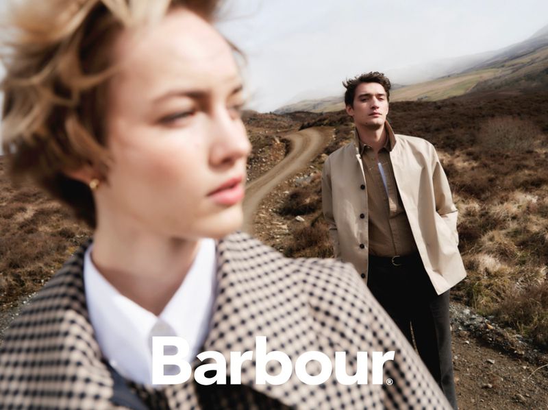Two models wearing Barbour clothing, posing on a dirt road winding through a hilly, grassy landscape, with the Barbour logo visible.