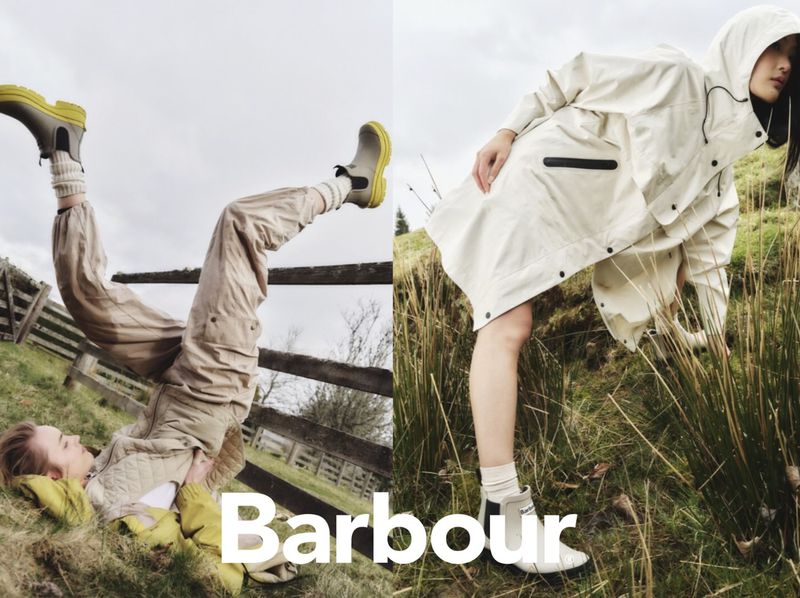 Two women wearing Barbour outdoor apparel in a field: one lying down with boots up, the other standing wearing a raincoat.