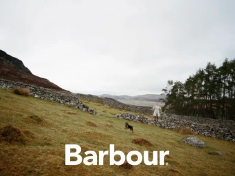 Model standing on a hillside near a drystone wall with a running dog under an overcast sky for Barbour S/S 25