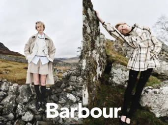 Model standing behind a stone wall in an outdoor rural landscape wearing a tan trench coat and white layered top for Barbour
